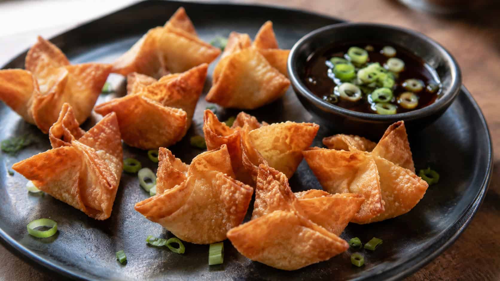crispy Peking duck wontons in a wide horizontal shot, arranged on a dark plate with a small dish of hoisin and scallion garnish, natural light, shallow depth of field, landscape orientation