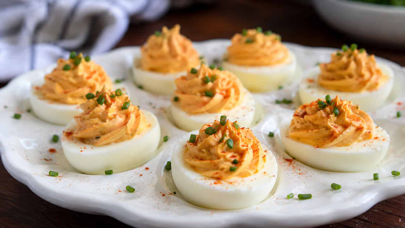 deviled eggs with creamy orange-tinted filling in a wide horizontal shot, arranged on a white deviled egg platter dusted with paprika and chives, natural light, shallow depth of field, landscape orientation
