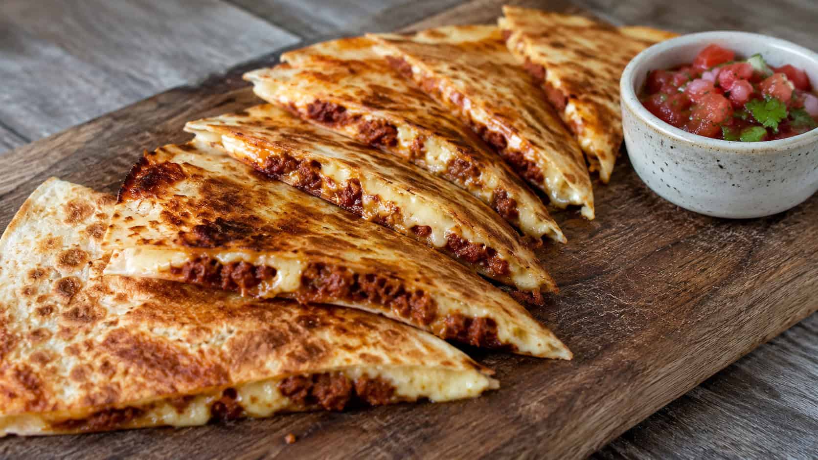 crispy golden quesadilla wedges with melty cheese and chorizo in a wide horizontal shot, fanned out on a wooden board with a small bowl of salsa, natural light, shallow depth of field, landscape orientation