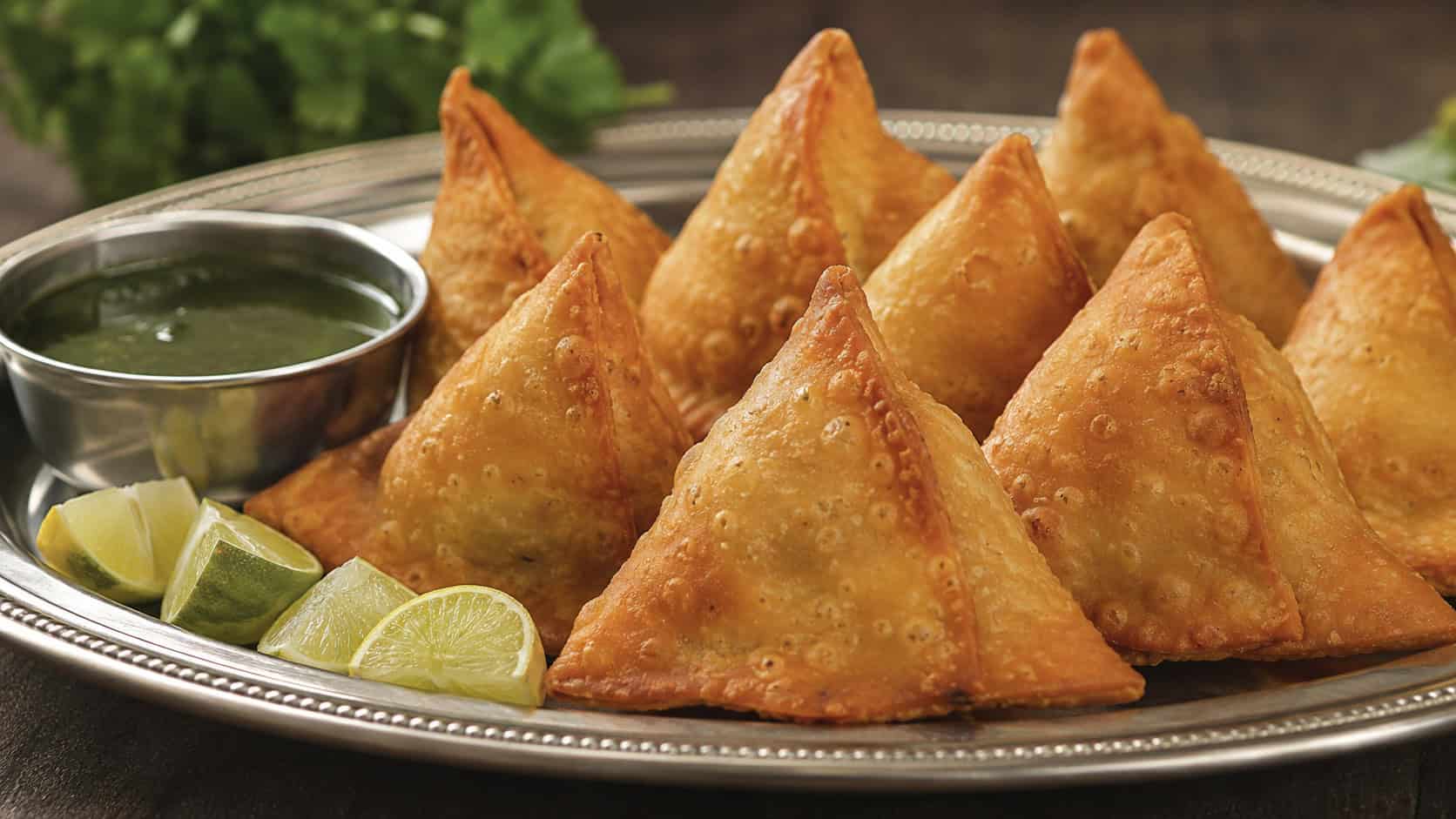 crispy golden triangular samosas in a wide horizontal shot, piled on a metal platter with a small bowl of green chutney and lime wedges, natural light, shallow depth of field, landscape orientation