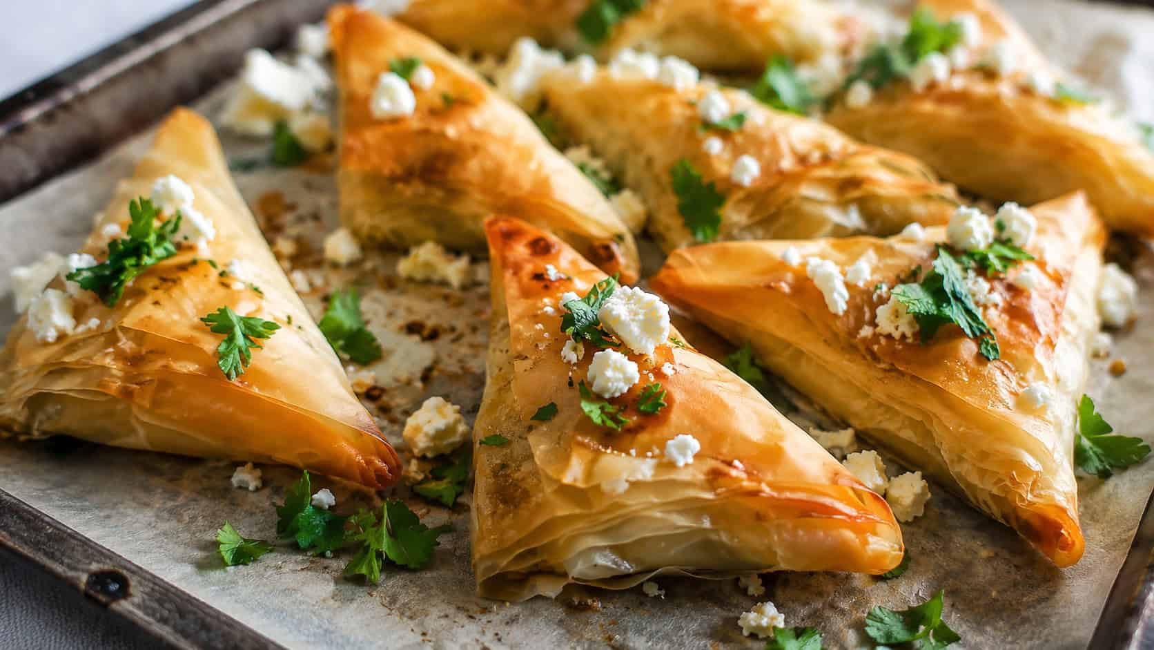 crisp golden phyllo triangles in a wide horizontal shot, arranged on parchment-lined tray with a scatter of fresh parsley and crumbled feta, natural light, shallow depth of field, landscape orientation
