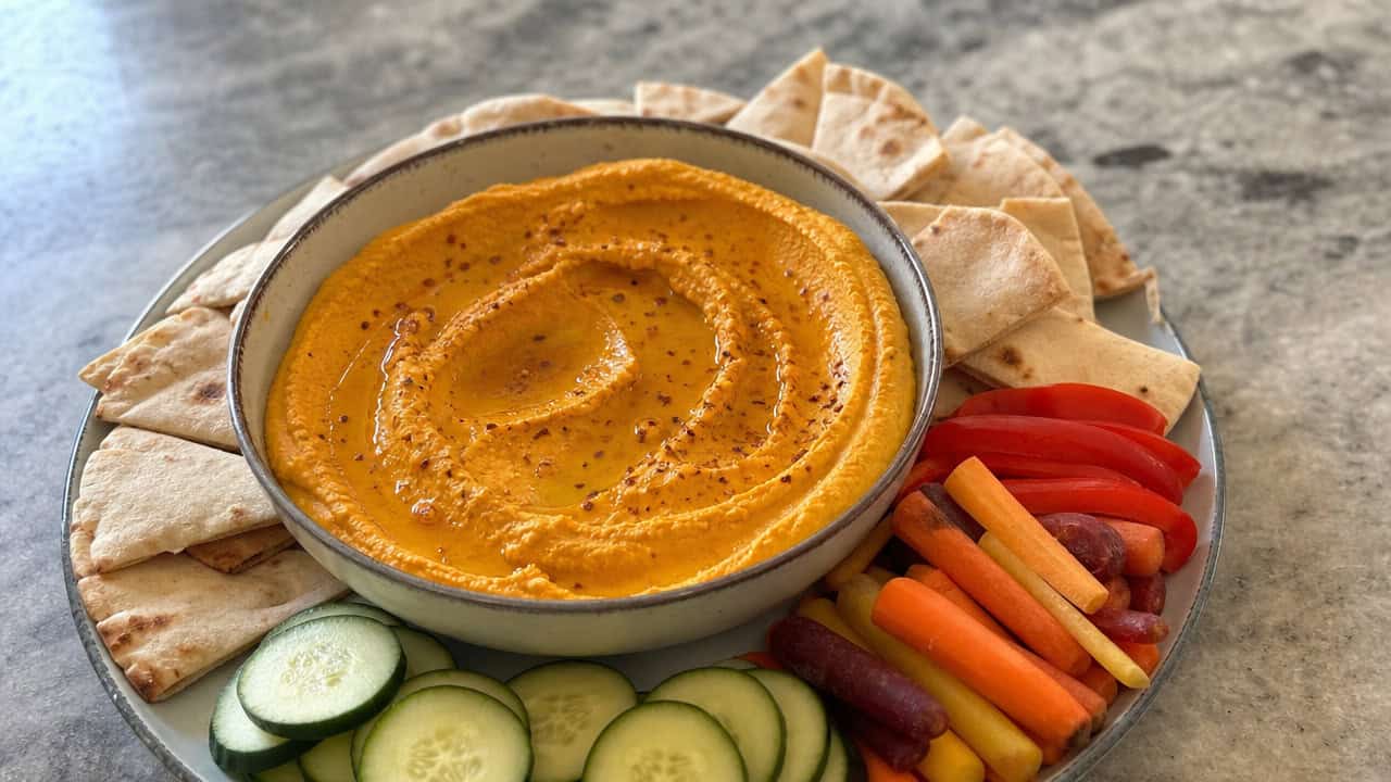 a bowl of creamy orange pumpkin hummus swirled and drizzled with olive oil in a wide horizontal shot, surrounded by pita wedges and crunchy vegetables on a stone countertop, natural light, shallow depth of field, landscape orientation