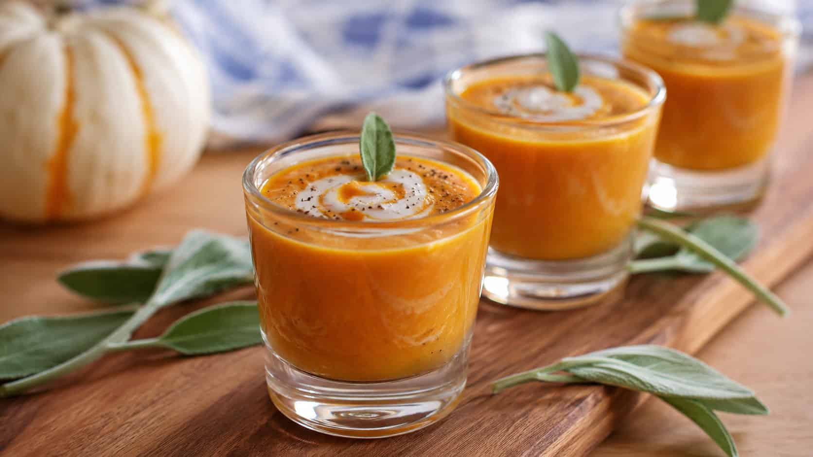 small glass shot glasses filled with creamy orange pumpkin soup swirled with cream in a wide horizontal shot, lined up on a wooden tray with sage leaves, natural light, shallow depth of field, landscape orientation
