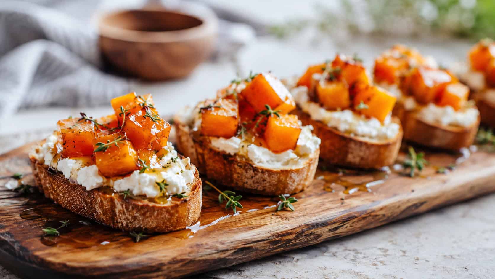 crostini topped with creamy white goat cheese and honeyed orange pumpkin in a wide horizontal shot, arranged in a row on a long wooden board drizzled with honey and thyme, natural light, shallow depth of field, landscape orientation