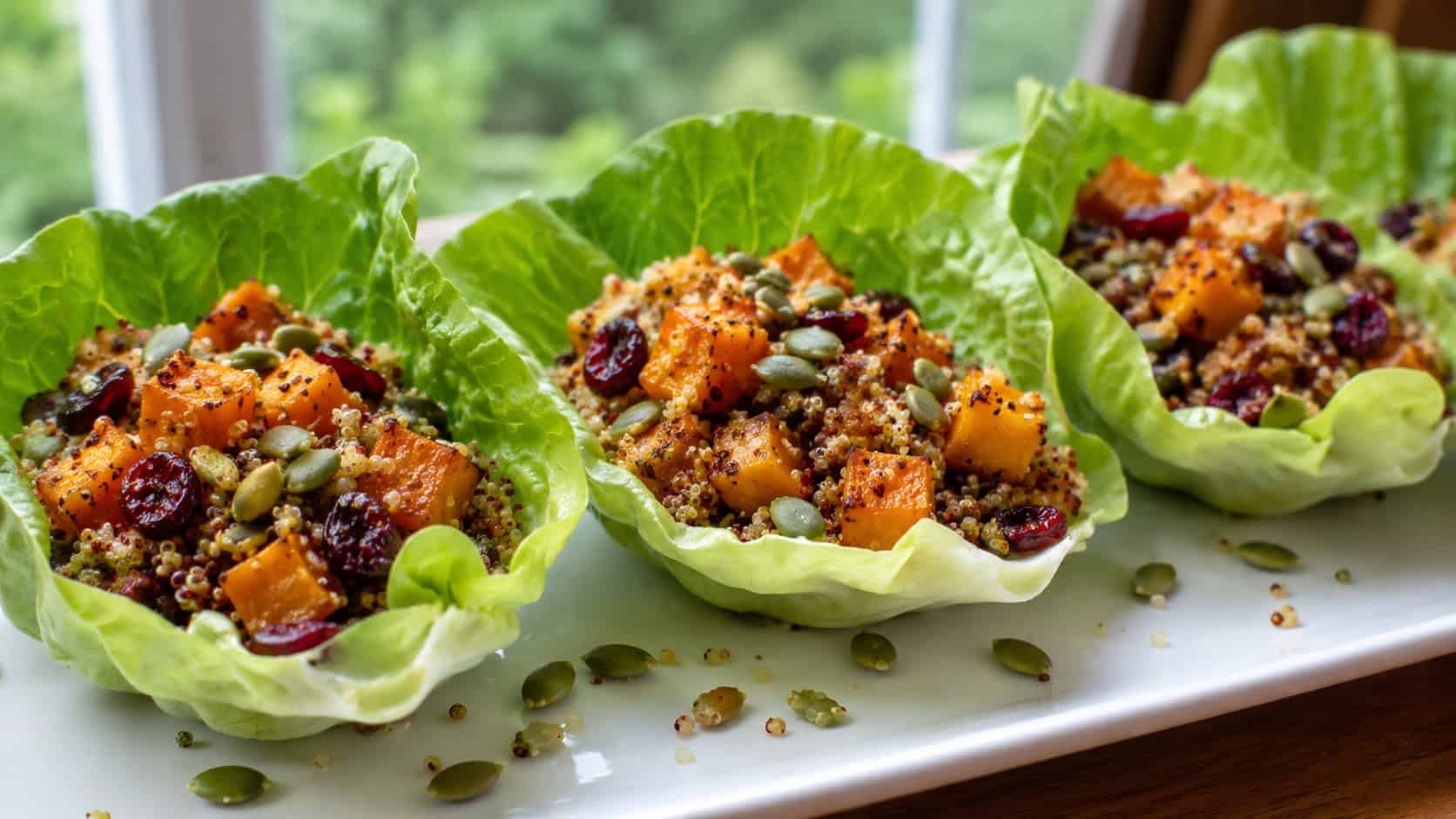 crisp green lettuce cups filled with quinoa, roasted pumpkin cubes, and cranberries in a wide horizontal shot, arranged on a white platter sprinkled with pumpkin seeds, natural light, shallow depth of field, landscape orientation