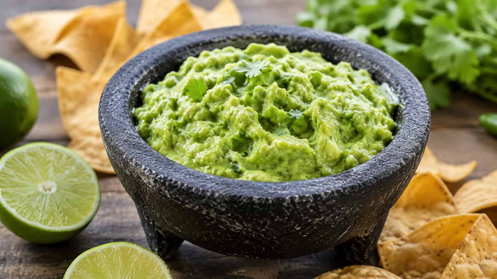 chunky green guacamole in a rustic stone molcajete in a wide horizontal composition, flecks of jalapeño and cilantro, lime halves and tortilla chips scattered around, bright natural light, shallow depth of field, landscape orientation