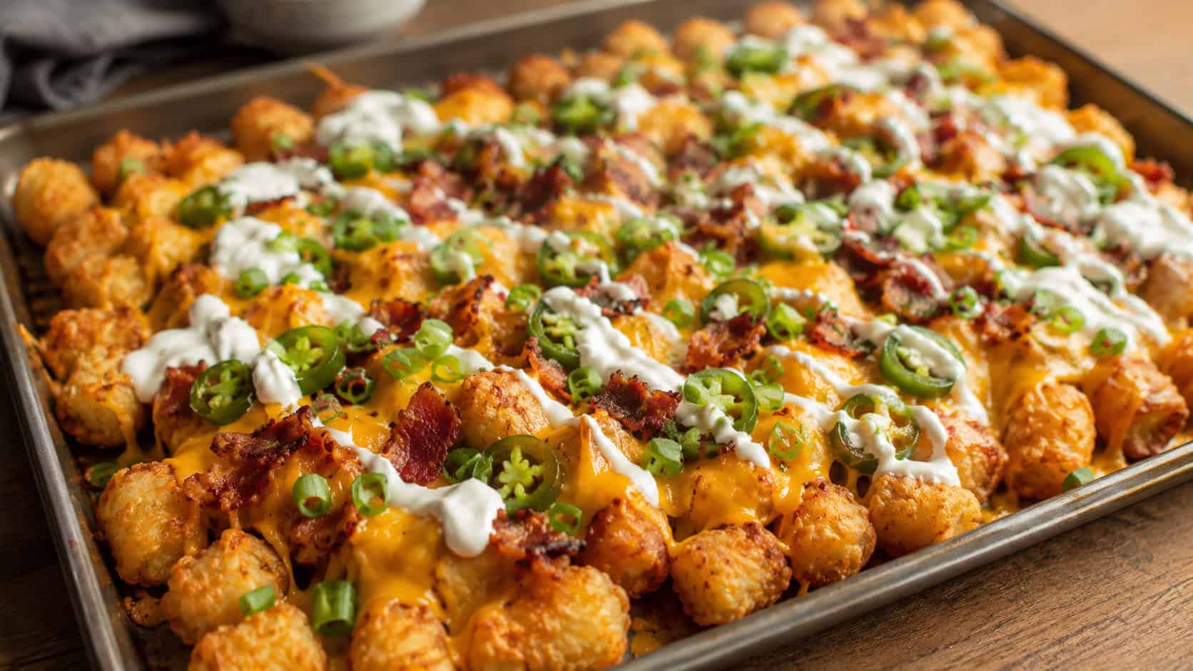 loaded tater tots on a sheet pan in a wide horizontal composition, melted cheddar and jalapeño slices, a drizzle of sour cream and green onion, crispy golden tots, warm natural light, shallow depth of field, landscape orientation