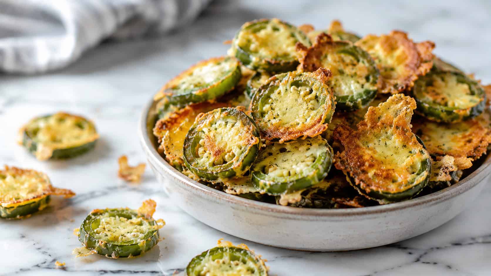 crispy baked jalapeño chips piled in a small bowl in a wide horizontal composition, golden parmesan edges, a few chips scattered on a marble counter, bright natural light, shallow depth of field, landscape orientation