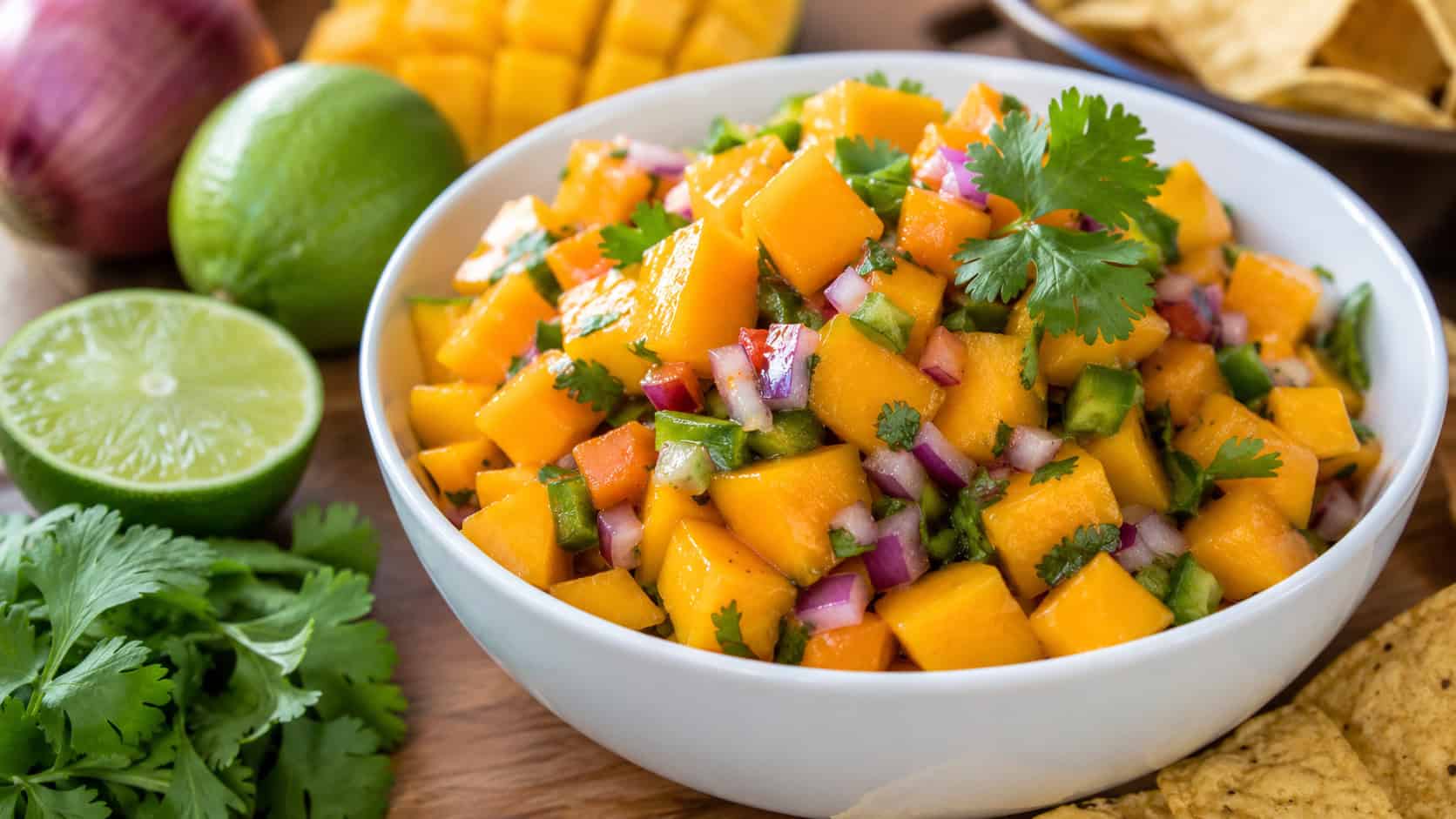 bright mango salsa in a white bowl in a wide horizontal composition, vivid orange mango with green jalapeño and red onion, lime halves and tortilla chips around it, fresh cilantro, natural light, shallow depth of field, landscape orientation