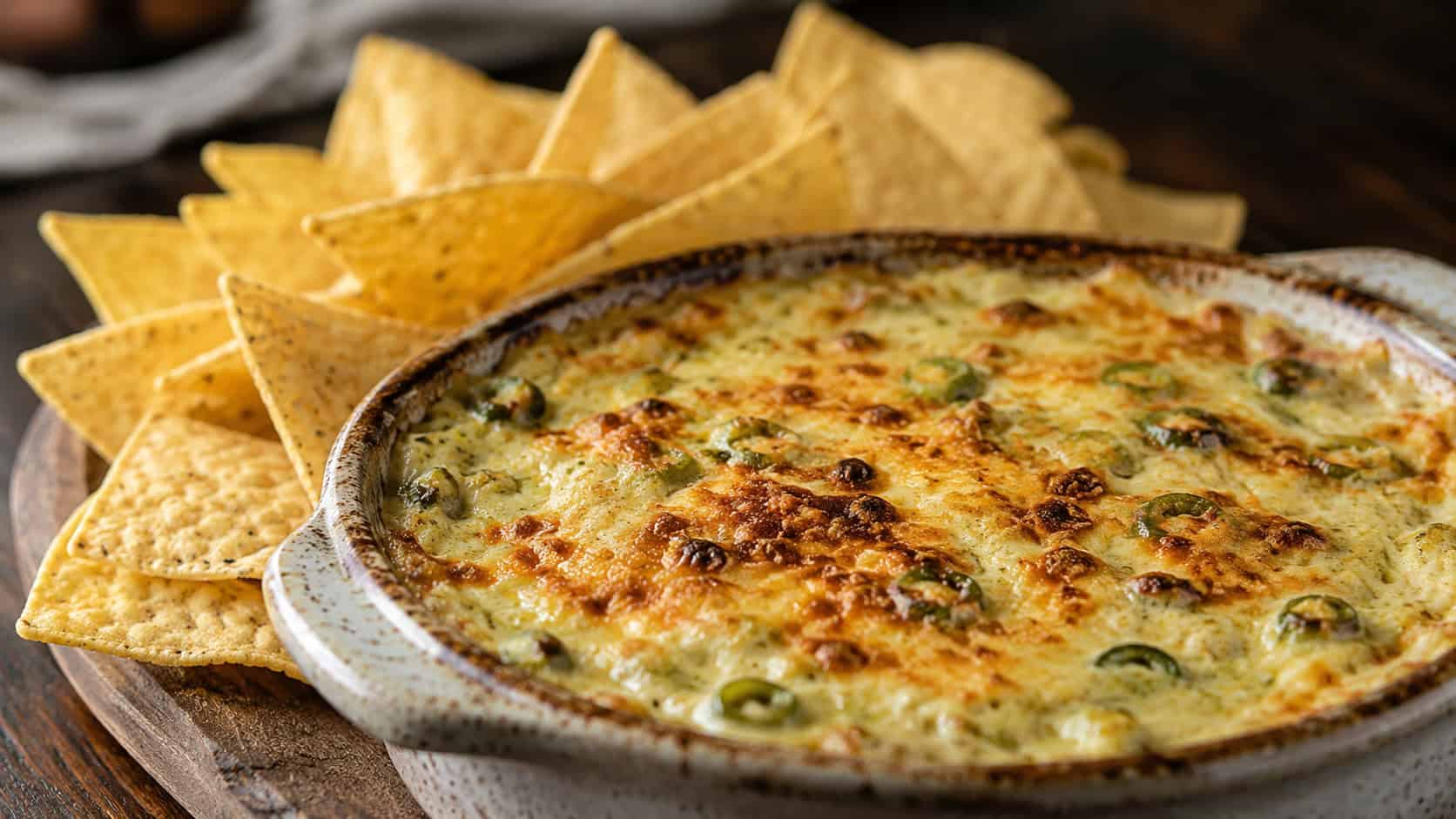 a bubbling golden artichoke jalapeño dip in a rustic baking dish in a wide horizontal composition, browned cheesy top, tortilla chips fanned around it, dark wooden table, warm natural light, shallow depth of field, landscape orientation