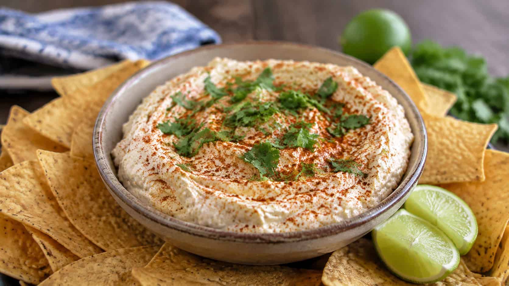 a bowl of whipped chili lime goat cheese spread topped with cilantro and a dusting of chili, surrounded by tortilla chips and lime wedges, in a wide horizontal shot, natural light, shallow depth of field, landscape orientation