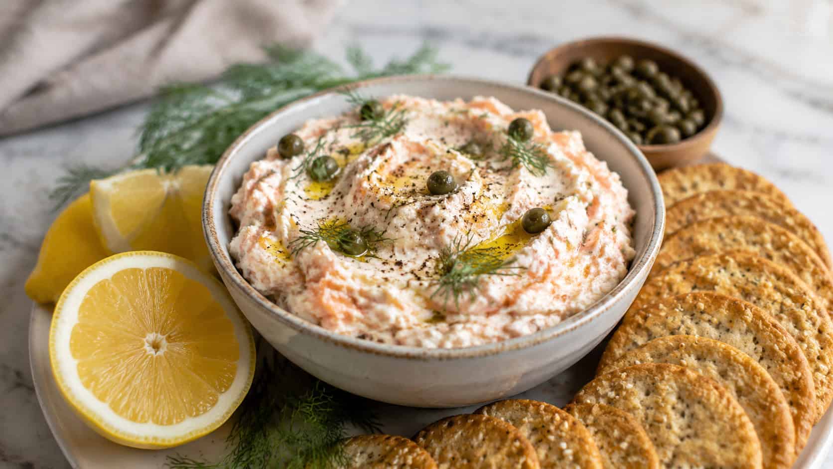 a bowl of pink-flecked smoked salmon goat cheese spread topped with dill and capers, surrounded by crackers and lemon, in a wide horizontal shot on a marble surface, natural light, shallow depth of field, landscape orientation