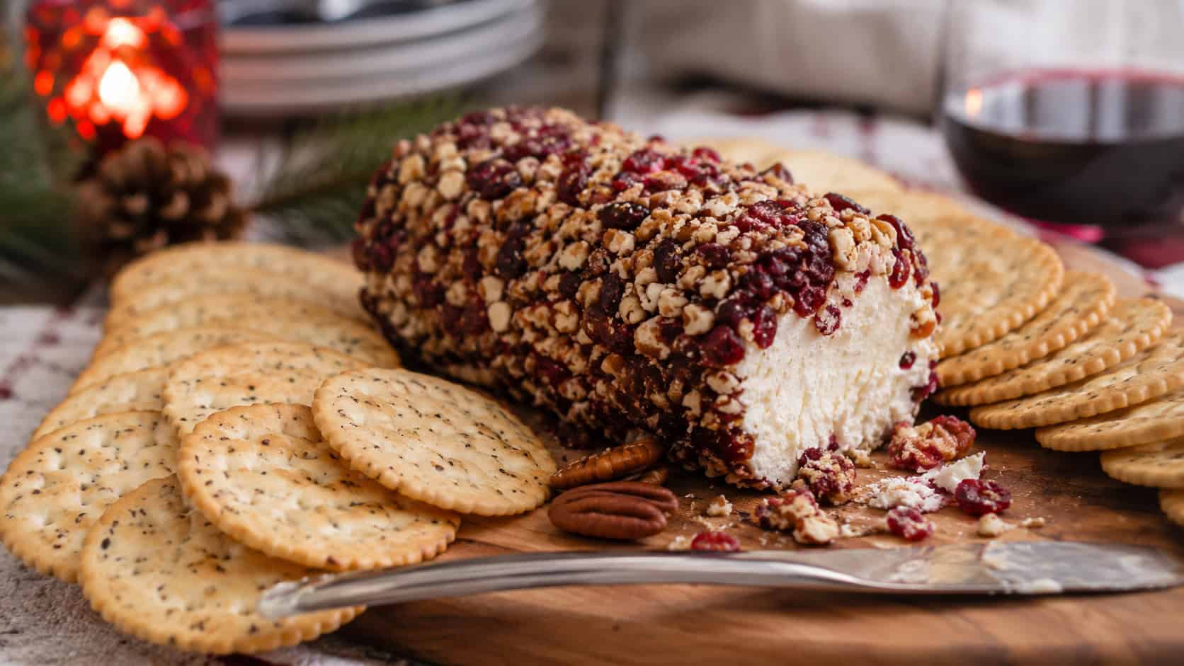 a goat cheese log coated in red cranberries and chopped pecans on a wooden board surrounded by crackers, in a wide horizontal shot on a holiday table, natural light, shallow depth of field, landscape orientation