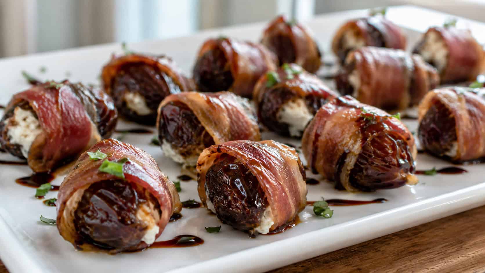 prosciutto-wrapped dates stuffed with goat cheese, crisp and glistening, arranged on a white platter with a balsamic drizzle, in a wide horizontal shot, natural light, shallow depth of field, landscape orientation