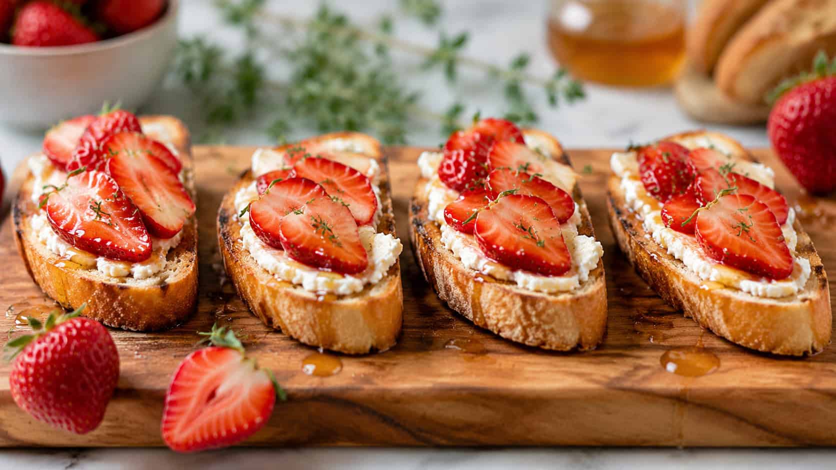 strawberry goat cheese toasts on crisp baguette slices drizzled with honey and thyme, arranged in a wide horizontal row on a wooden board, fresh strawberries scattered nearby, bright natural light, shallow depth of field, landscape orientation