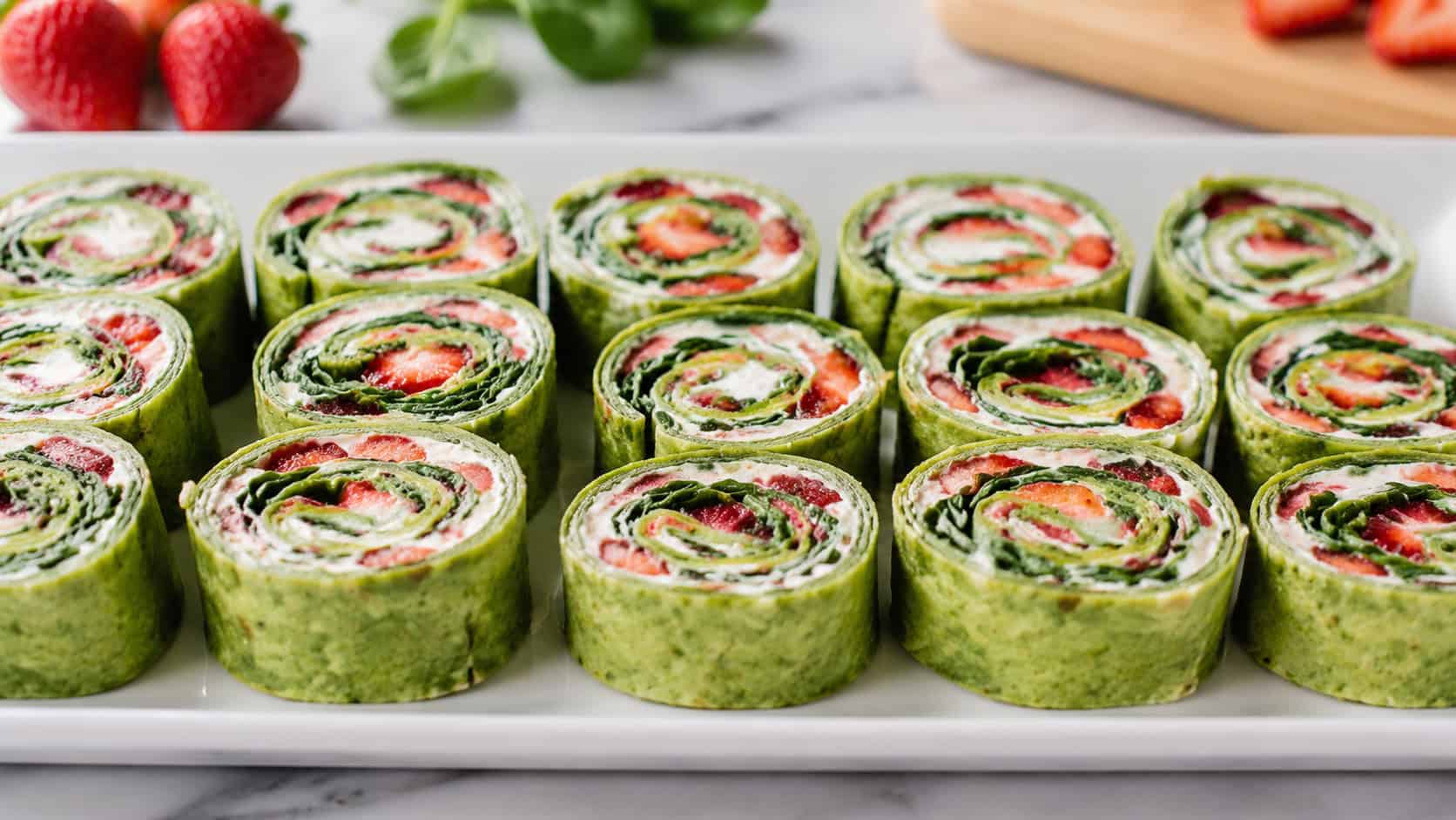 strawberry spinach pinwheels showing colorful spirals of red and green, arranged cut-side up in a wide horizontal row on a white platter, bright natural light, shallow depth of field, landscape orientation