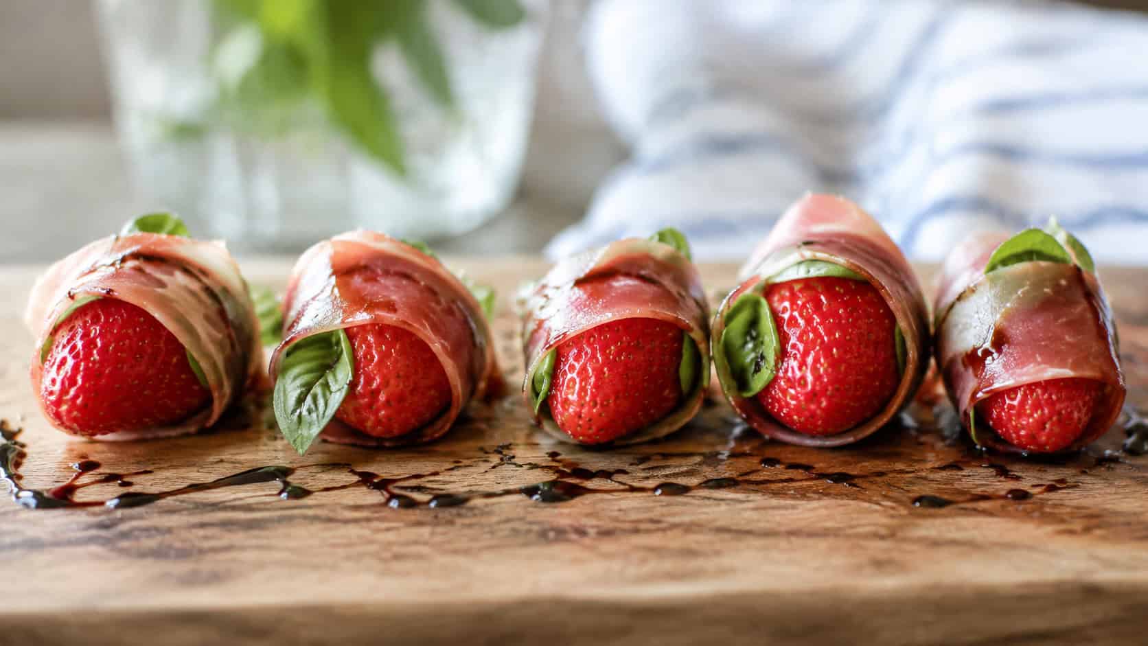 prosciutto-wrapped strawberries with peeks of basil, arranged in a wide horizontal row on a wooden board drizzled with balsamic, soft natural light, shallow depth of field, landscape orientation