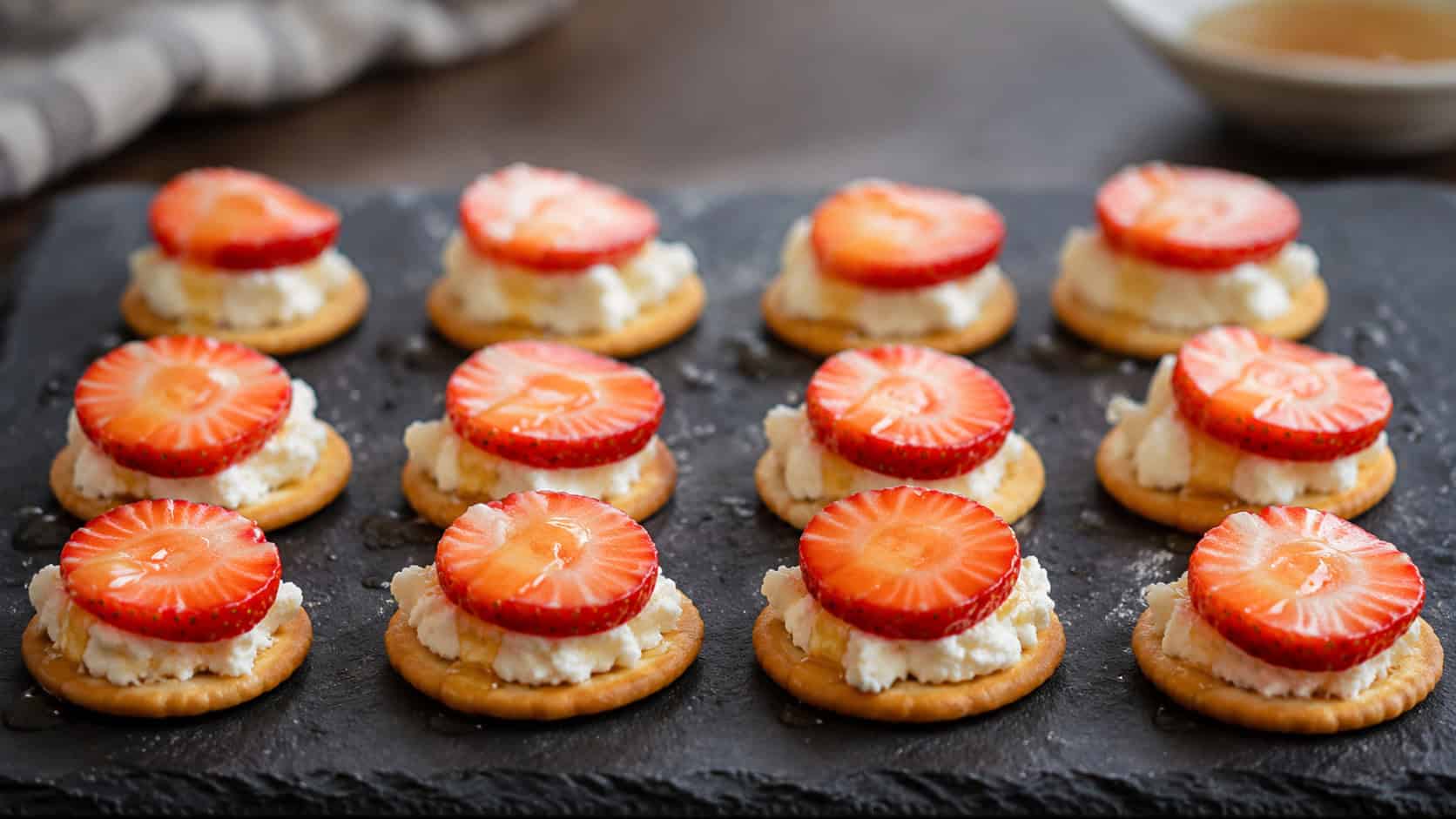 round crackers topped with creamy goat cheese and a strawberry slice drizzled with honey, arranged in a wide horizontal grid on a slate board, soft natural light, shallow depth of field, landscape orientation