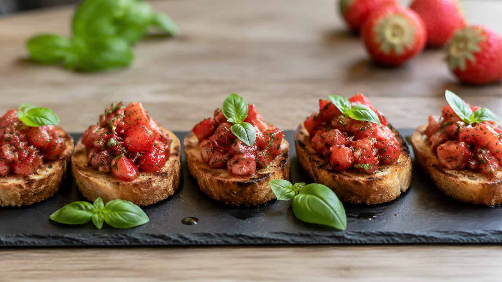 strawberry basil bruschetta with chunky red relish piled on toasted bread, arranged in a wide horizontal line on a slate board with fresh basil leaves, soft natural light, shallow depth of field, landscape orientation