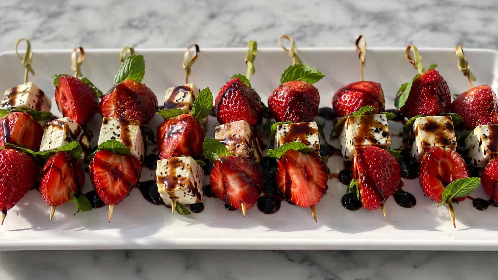strawberry and feta skewers drizzled with dark balsamic glaze, arranged in a wide horizontal row on a white platter with mint leaves, bright natural light, shallow depth of field, landscape orientation