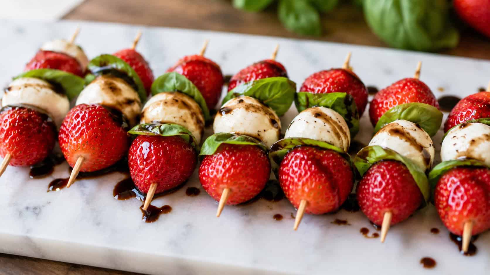 strawberry caprese skewers with red strawberries, white mozzarella, and green basil drizzled with balsamic glaze, arranged in a wide horizontal line on a marble board, bright natural light, shallow depth of field, landscape orientation