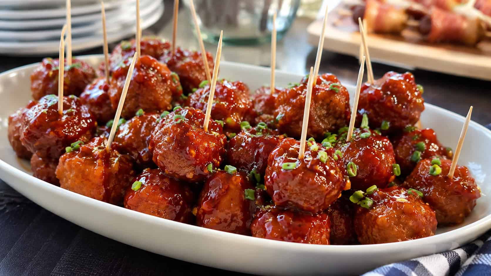 glossy sweet-and-spicy sausage meatballs piled in a shallow serving dish with toothpicks, in a wide horizontal shot, a glistening red glaze and chopped chives on top, on a party table, natural light, shallow depth of field, landscape orientation