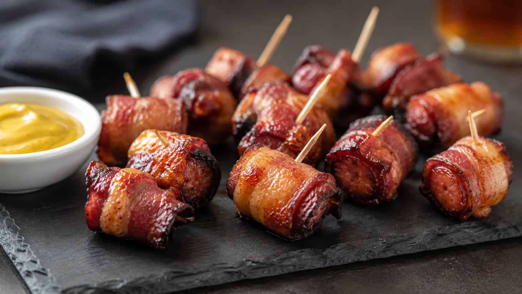 crispy bacon-wrapped smoked sausage bites with caramelized brown sugar edges on a slate serving board, in a wide horizontal shot, toothpicks and a small dish of mustard nearby, on a dark table, natural light, shallow depth of field, landscape orientation