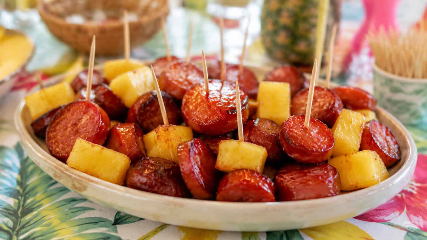 glossy pineapple-glazed smoked sausage coins in a shallow ceramic dish with toothpicks and pineapple chunks, in a wide horizontal shot, on a bright party table, natural light, shallow depth of field, landscape orientation