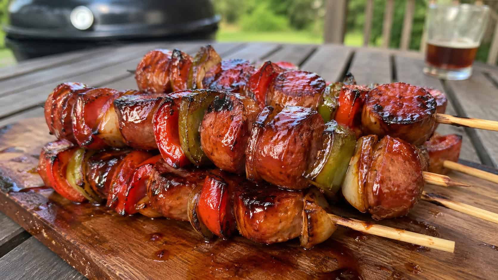 grilled smoked sausage skewers with charred peppers and onions glazed in barbecue sauce on a wooden board, in a wide horizontal shot, light grill marks visible, on an outdoor table, natural light, shallow depth of field, landscape orientation