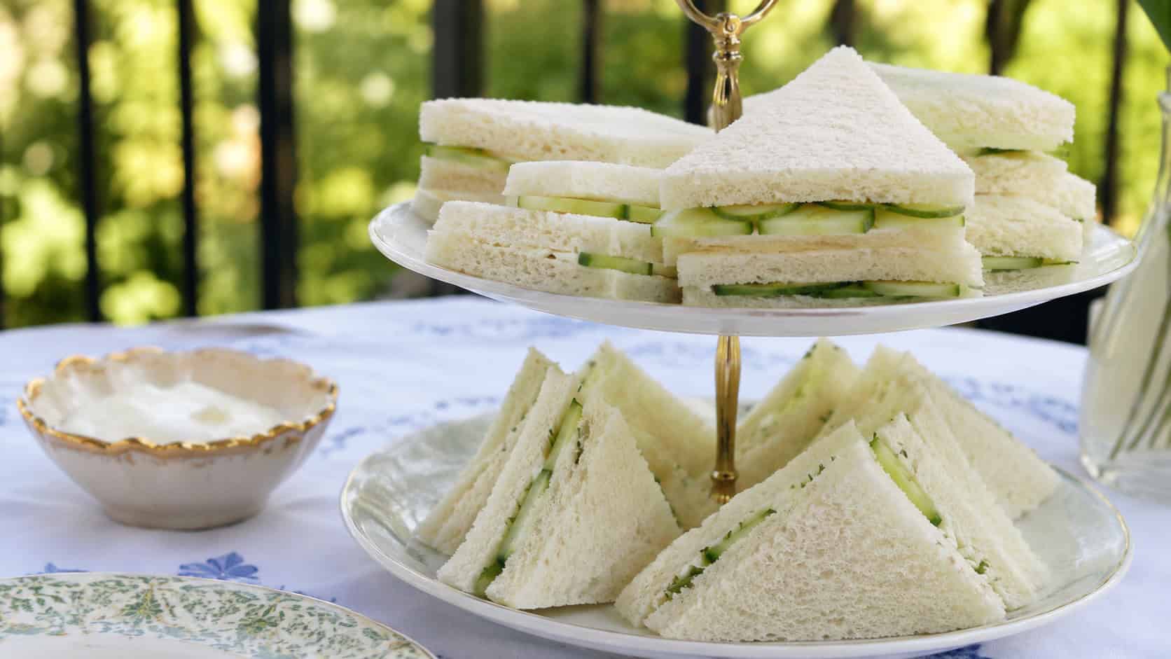 dainty cucumber sandwiches cut into triangles with green cucumber peeking out, stacked on a tiered tray, in a wide horizontal shot on a bright outdoor table, natural light, shallow depth of field, landscape orientation
