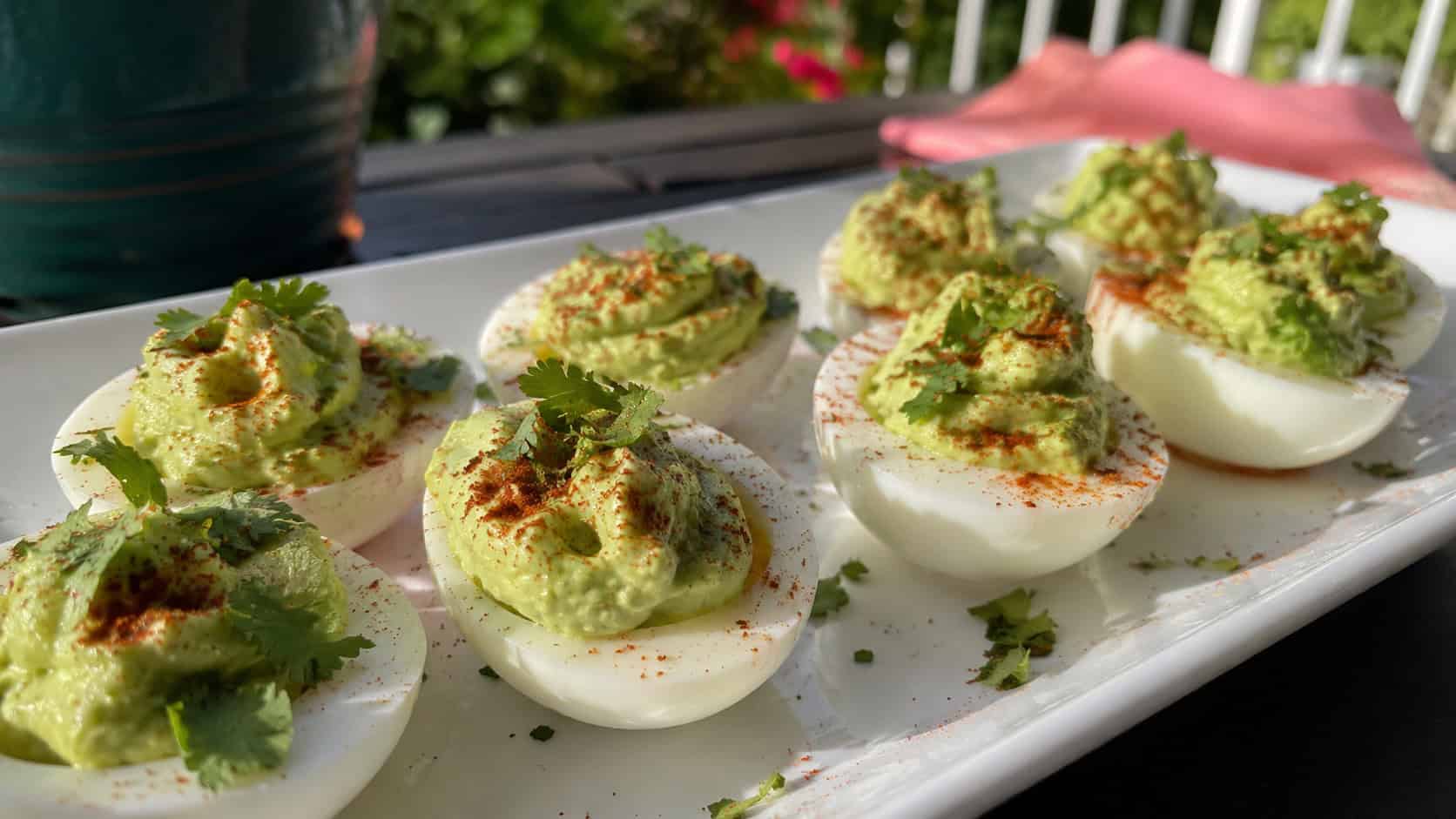deviled eggs with creamy green avocado filling dusted with paprika and cilantro on a white platter, in a wide horizontal shot on a sunny outdoor table, natural light, shallow depth of field, landscape orientation