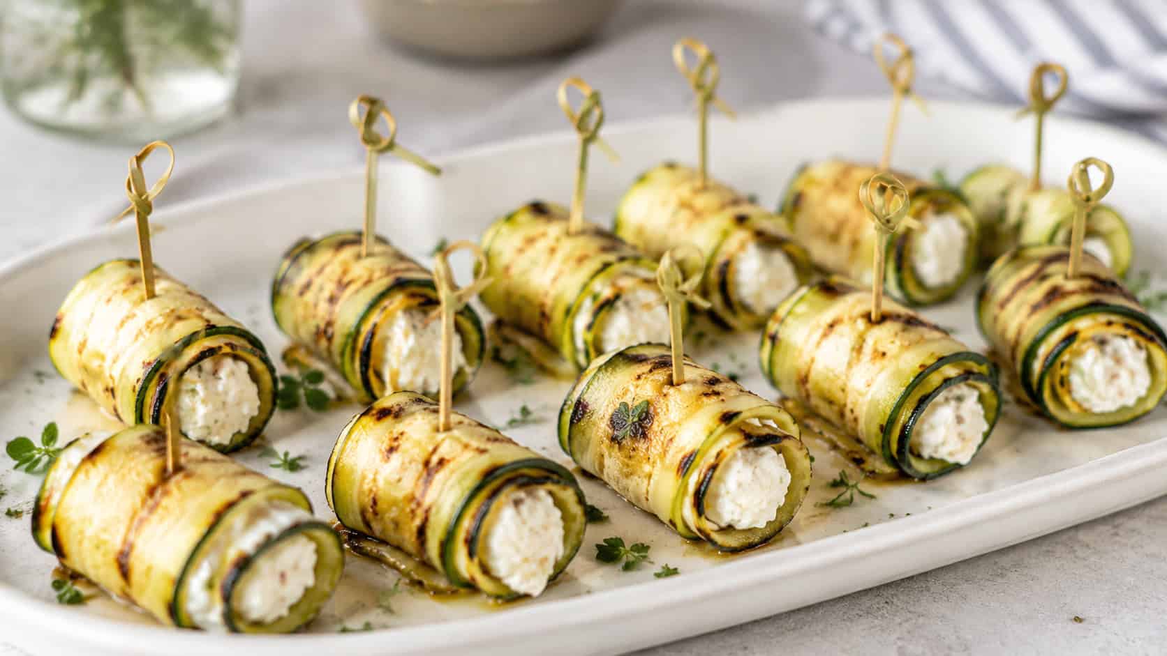 grilled zucchini rolls with char marks wrapped around creamy white filling, secured with picks on a platter, in a wide horizontal shot on a bright table, natural light, shallow depth of field, landscape orientation