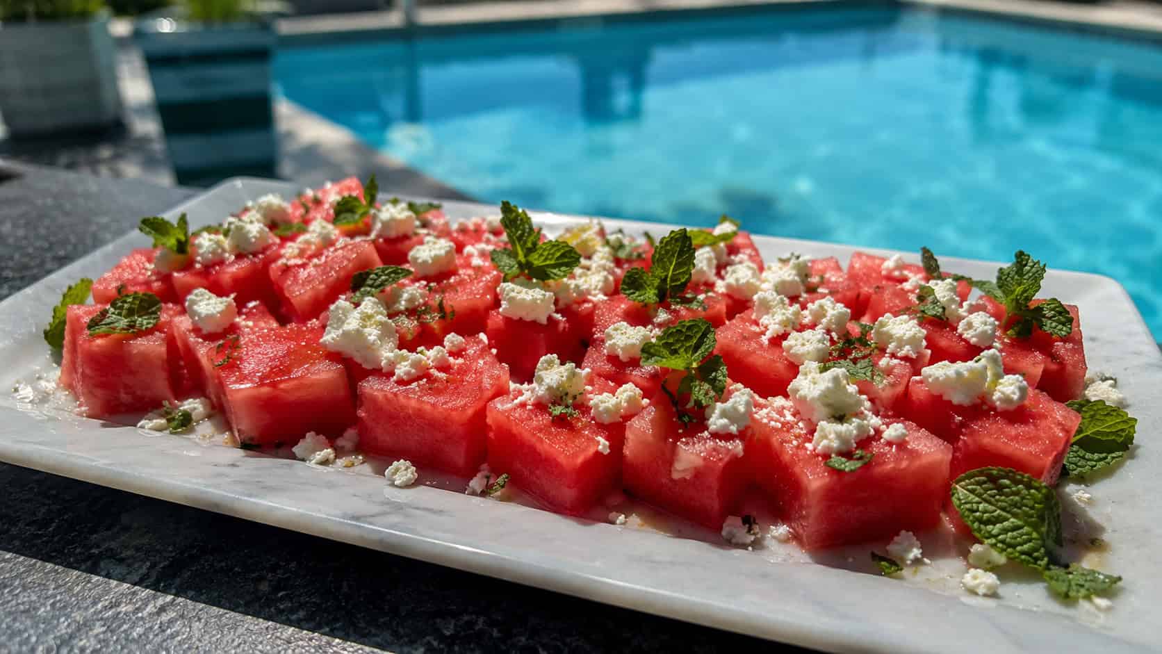 bright pink watermelon cubes topped with white feta and green mint leaves on a chilled platter, in a wide horizontal shot beside a poolside table, natural light, shallow depth of field, landscape orientation