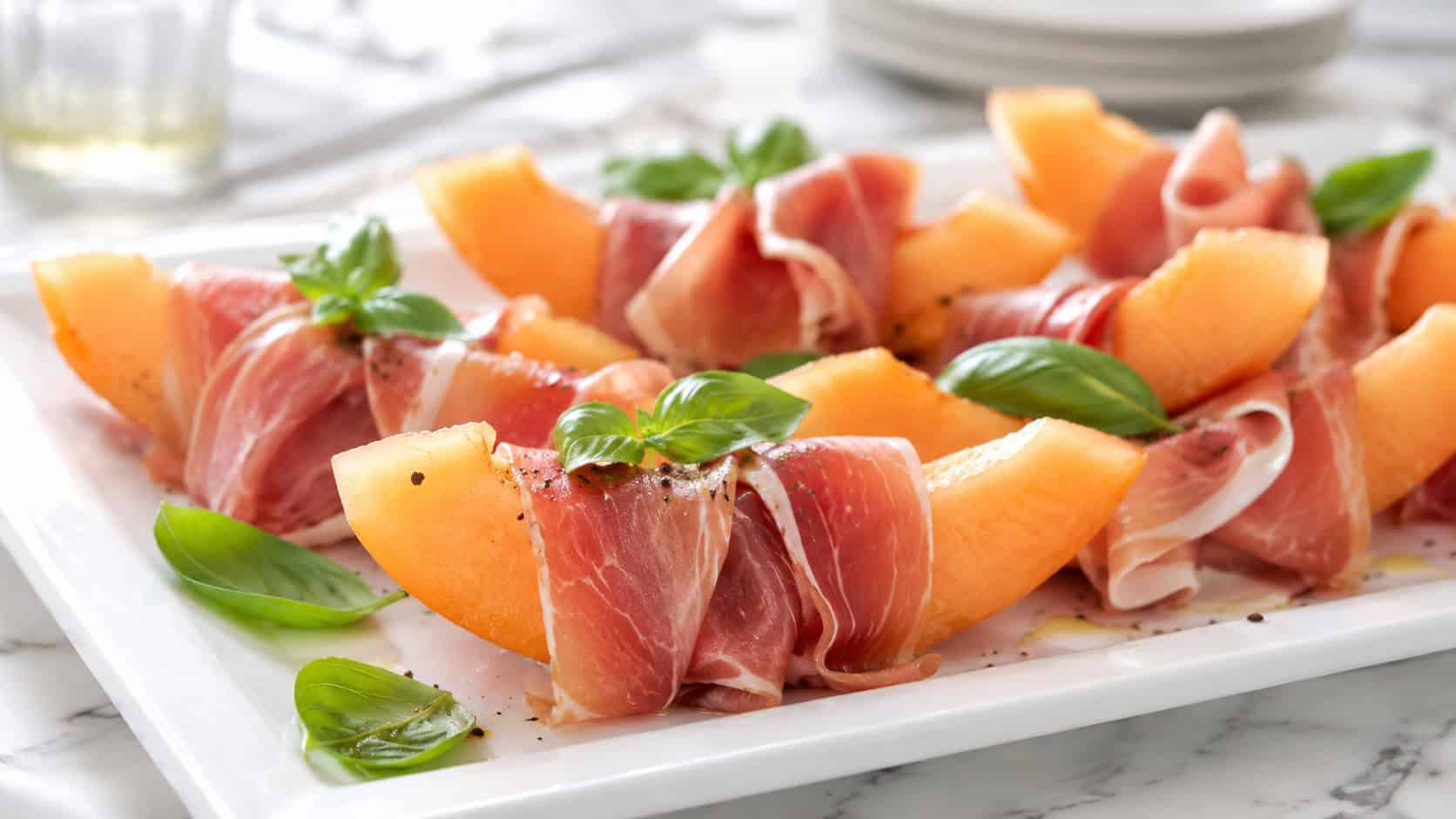 orange cantaloupe wedges wrapped in ruffled pink prosciutto with basil leaves on a white platter, in a wide horizontal shot on a bright table, natural light, shallow depth of field, landscape orientation