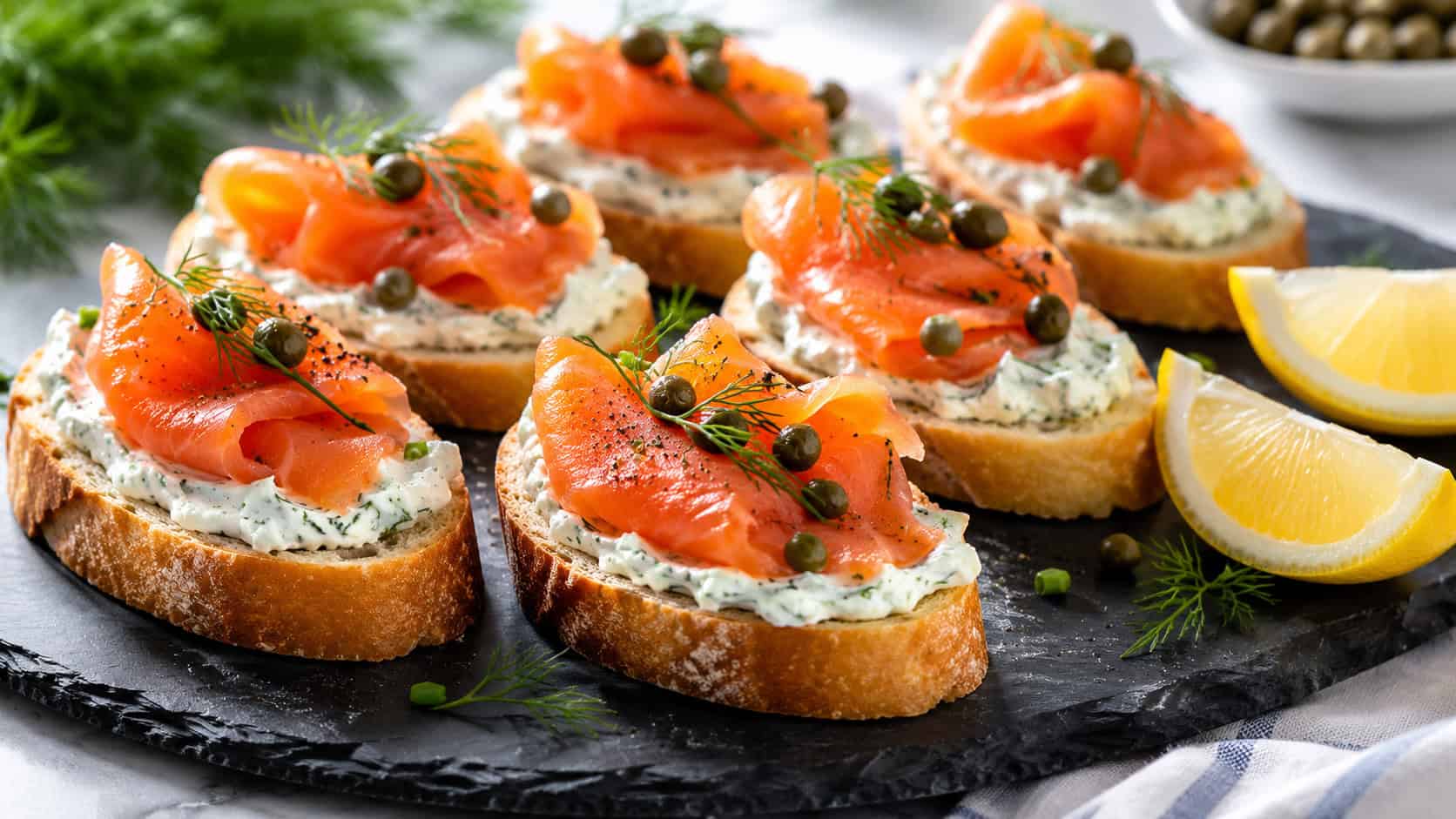 elegant baguette canapes topped with herbed cream cheese, smoked salmon, and capers in a wide horizontal shot, arranged on a slate platter with lemon wedges, bright natural light, shallow depth of field, landscape orientation
