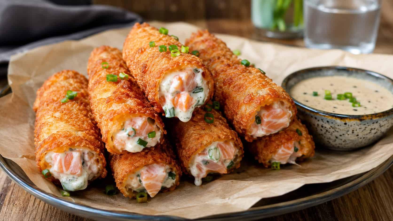 crispy golden fried salmon rolls with a creamy filling peeking out in a wide horizontal shot, on a plate lined with parchment over a wooden table with a dipping sauce, warm natural light, shallow depth of field, landscape orientation
