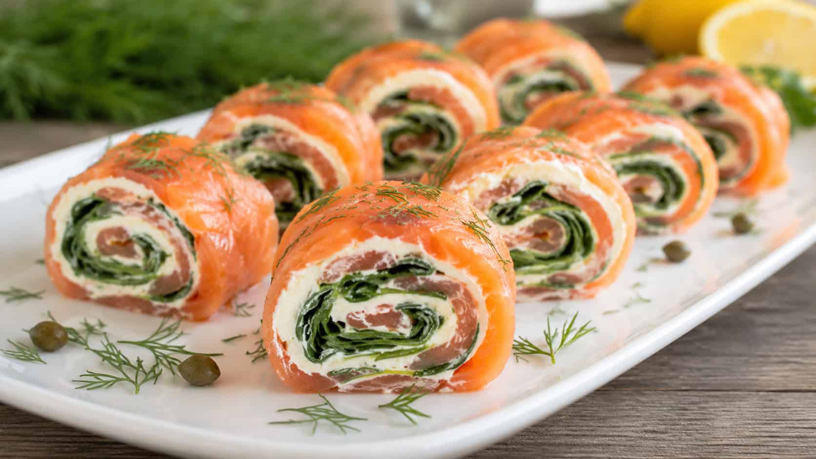smoked salmon pinwheels showing spiral layers of cream cheese, salmon, and spinach in a wide horizontal shot, arranged on a white platter over a wooden table, bright natural light, shallow depth of field, landscape orientation