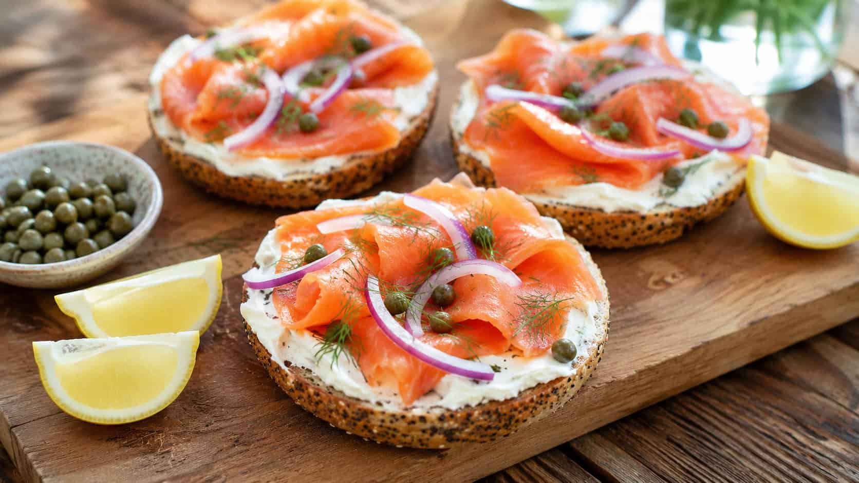open-faced bagels piled with cream cheese, smoked salmon, red onion, and capers in a wide horizontal shot, on a board over a wooden table with lemon wedges nearby, bright natural light, shallow depth of field, landscape orientation