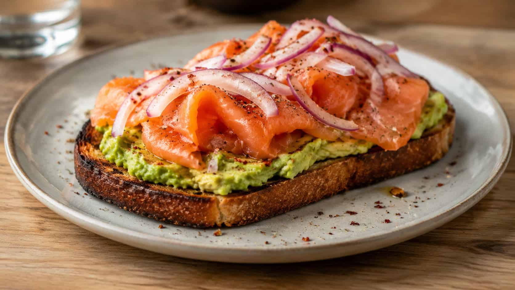 avocado toast topped with smoked salmon and red onion in a wide horizontal shot, on a plate over a wooden table with a sprinkle of red pepper flakes, bright natural light, shallow depth of field, landscape orientation