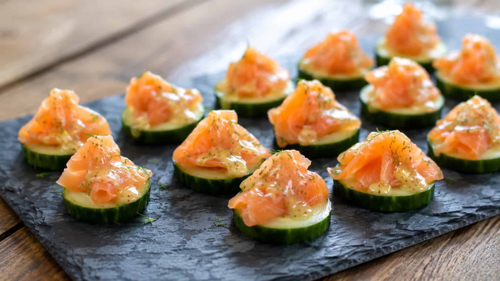 cucumber rounds topped with smoked salmon and a honey-dijon drizzle in a wide horizontal shot, arranged on a slate platter over a wooden table, bright natural light, shallow depth of field, landscape orientation