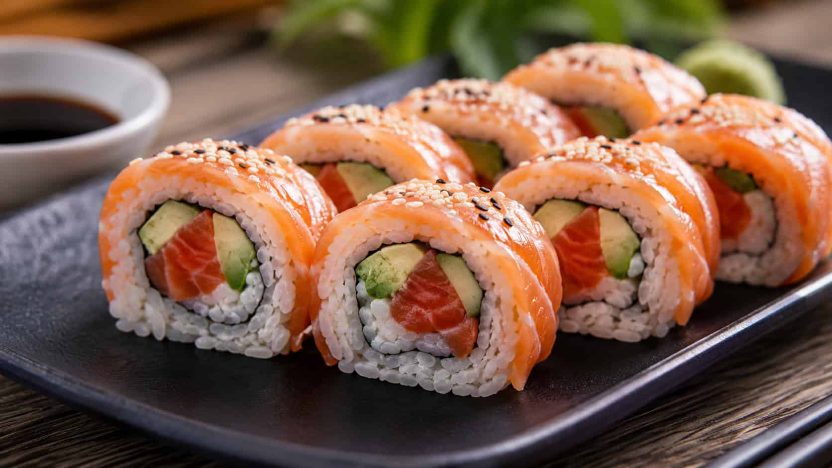 smoked salmon sushi rolls sliced into rounds showing rice, salmon, and avocado in a wide horizontal shot, arranged on a dark plate with soy sauce nearby, bright natural light, shallow depth of field, landscape orientation