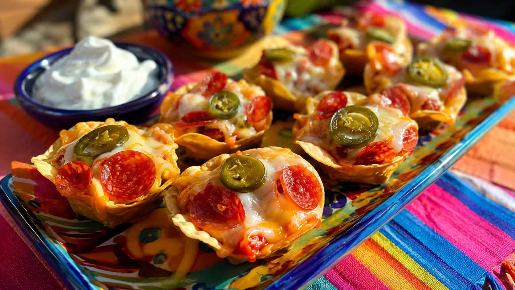 individual loaded pepperoni nacho bites with melted cheese and jalapeno on a platter in a wide horizontal shot, with a dollop of sour cream on a colorful table, warm bright light, shallow depth of field, landscape orientation
