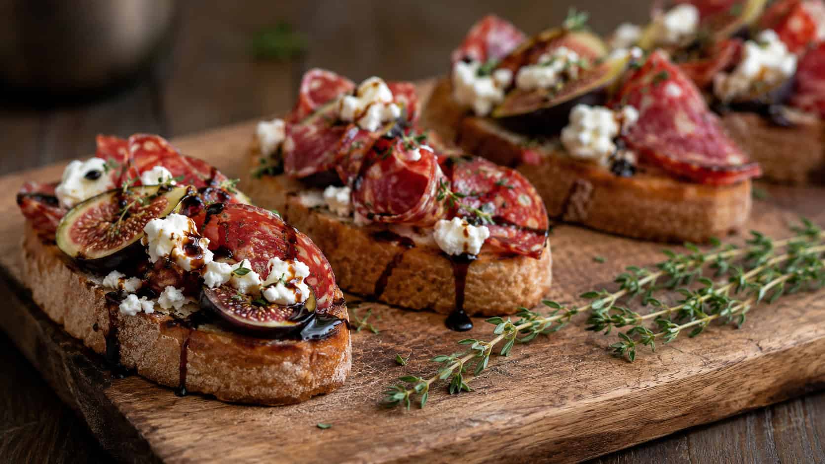 pepperoni and fig bruschetta topped with goat cheese and balsamic drizzle in a wide horizontal shot, lined on a wooden board with thyme sprigs, warm rustic light, shallow depth of field, landscape orientation
