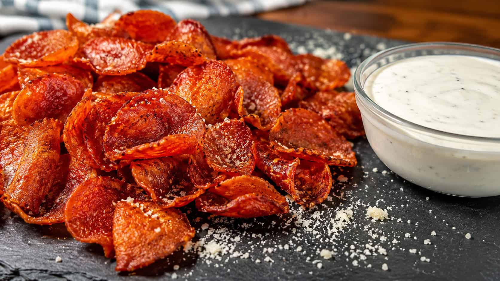 a pile of crispy curled pepperoni chips beside a bowl of ranch dip in a wide horizontal shot, on a slate board with a sprinkle of Parmesan, bright crisp light, shallow depth of field, landscape orientation