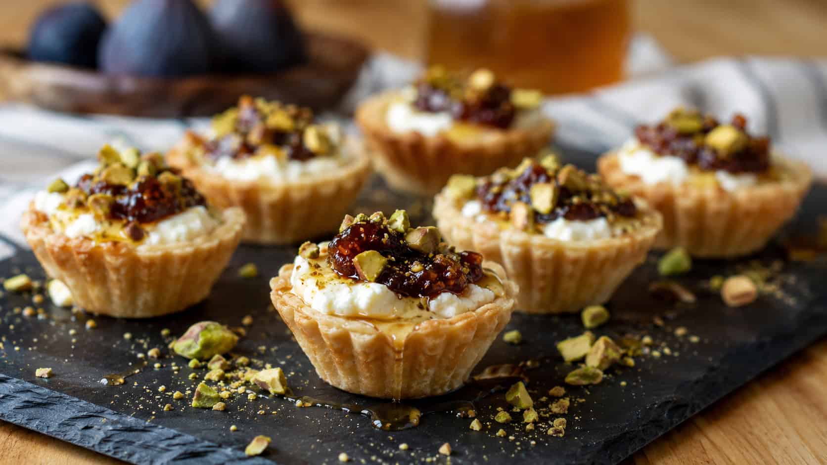 mini tartlets filled with creamy goat cheese and dark fig jam, drizzled with honey and topped with crushed pistachios, arranged on a slate board, in a wide horizontal shot, warm natural light, shallow depth of field, landscape orientation