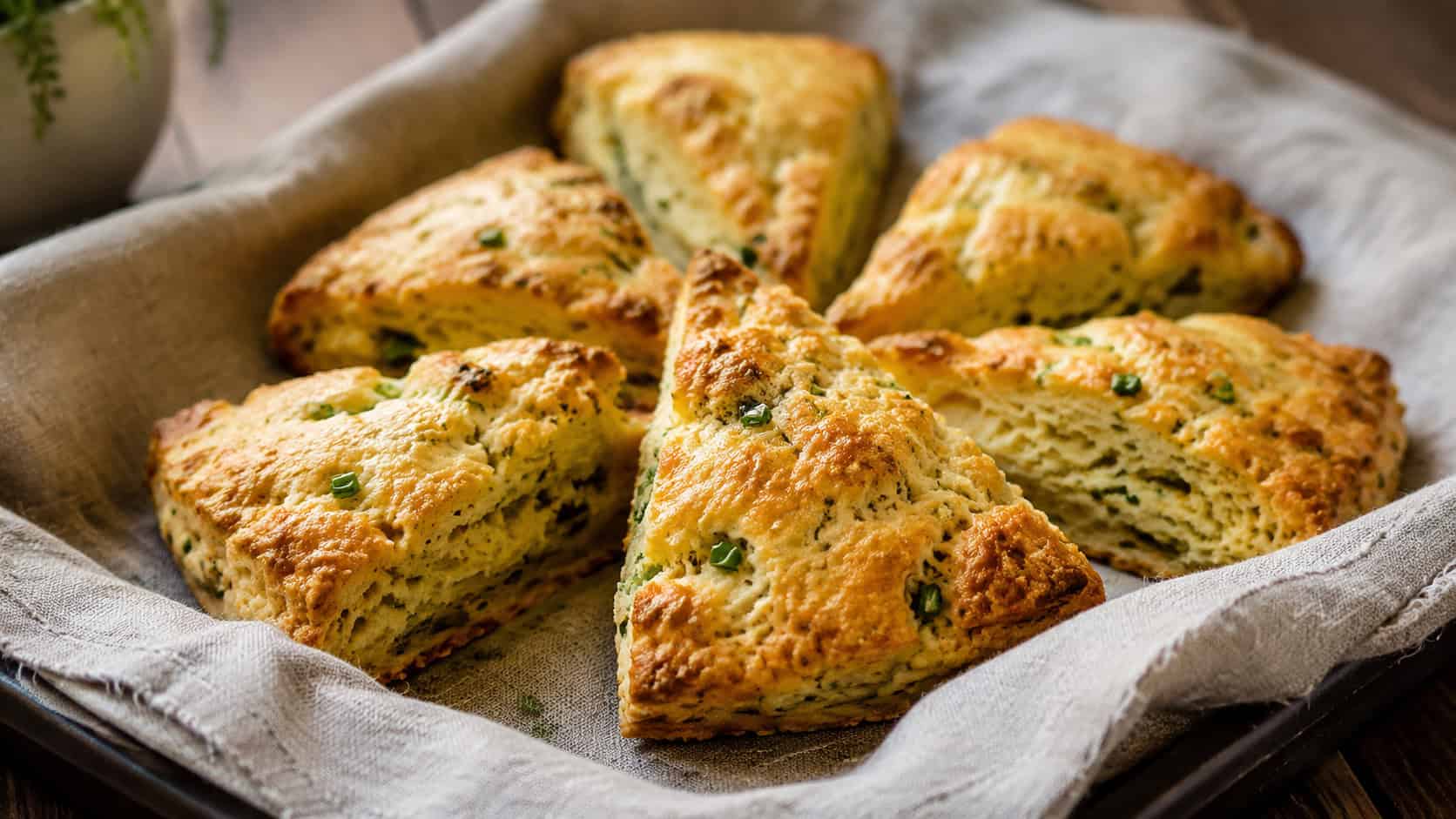 golden savory herb and garlic scones cut into wedges, flecked with green chives, arranged on a linen-lined tray, in a wide horizontal shot, soft natural light, shallow depth of field, landscape orientation