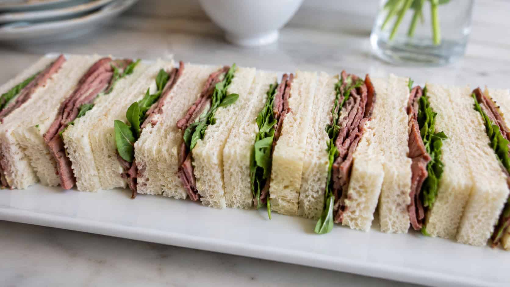roast beef tea sandwiches cut into fingers with a peek of arugula, arranged on a white rectangular platter, in a wide horizontal shot, soft natural light, shallow depth of field, landscape orientation