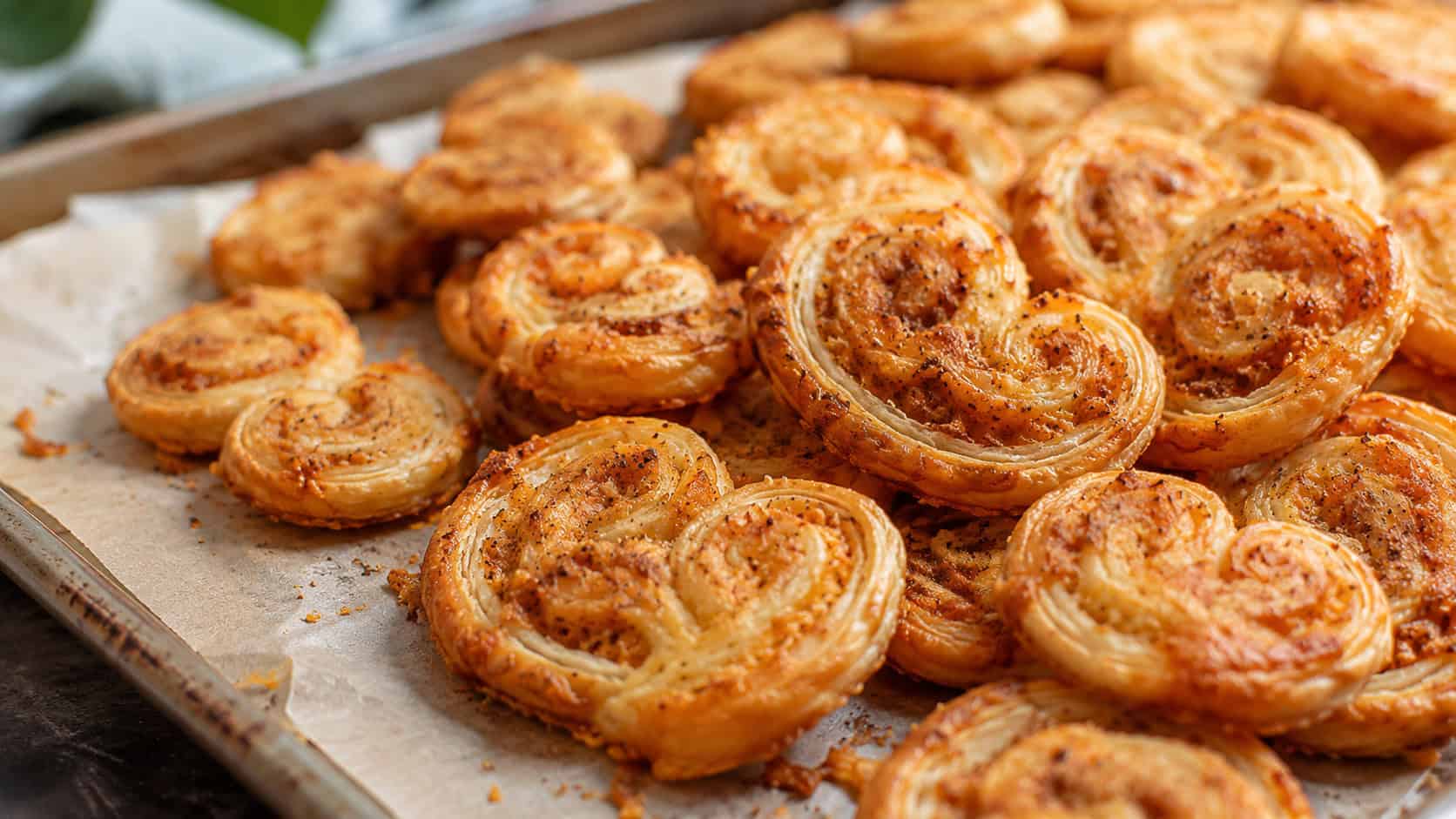 golden savory palmiers with visible spirals of Parmesan, piled on a parchment-lined tray, in a wide horizontal shot, warm natural light, shallow depth of field, landscape orientation