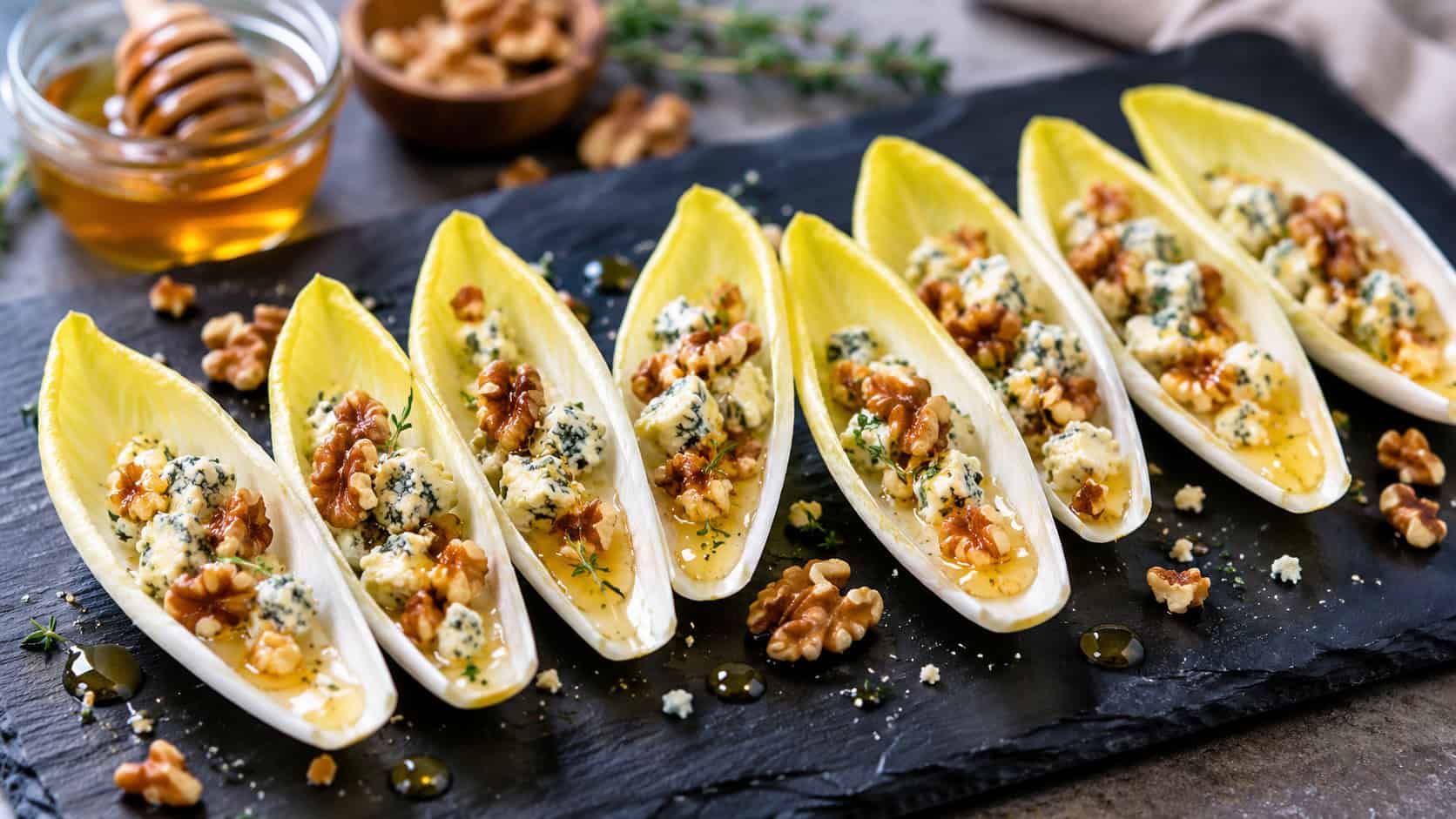 crisp endive leaves filled with crumbled blue cheese and walnuts drizzled with honey, fanned out on a dark slate board, in a wide horizontal shot, natural light, shallow depth of field, landscape orientation