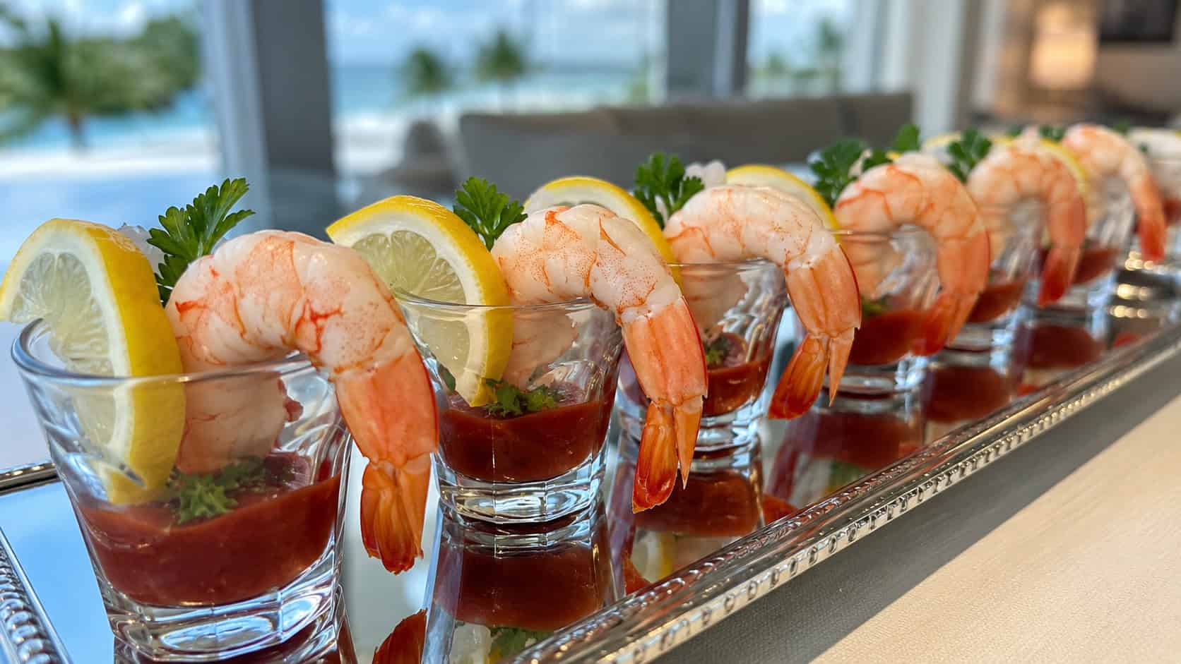 chilled shrimp cocktail served in small glass cups with red cocktail sauce and lemon wedges, lined up on a mirrored tray, in a wide horizontal shot, bright natural light, shallow depth of field, landscape orientation